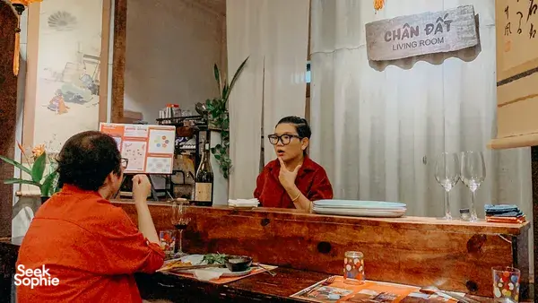 Host in a red robe welcomes guests at Chan Dat Living Room in Hoi An, with wine glasses and tasting materials on the table