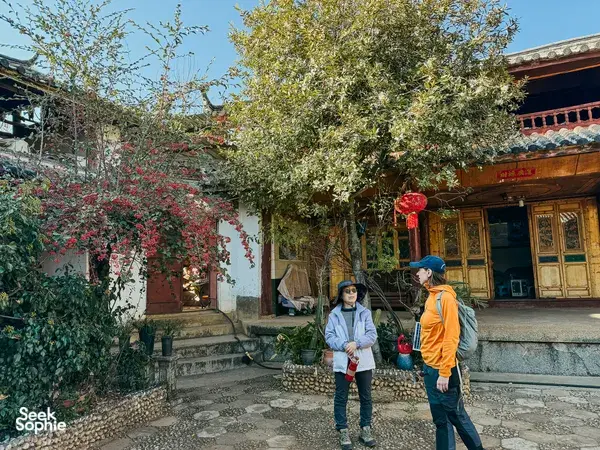 Two travellers walking a stone courtyard in Lijiang, passing flowering trees beside a traditional wooden door during the trek