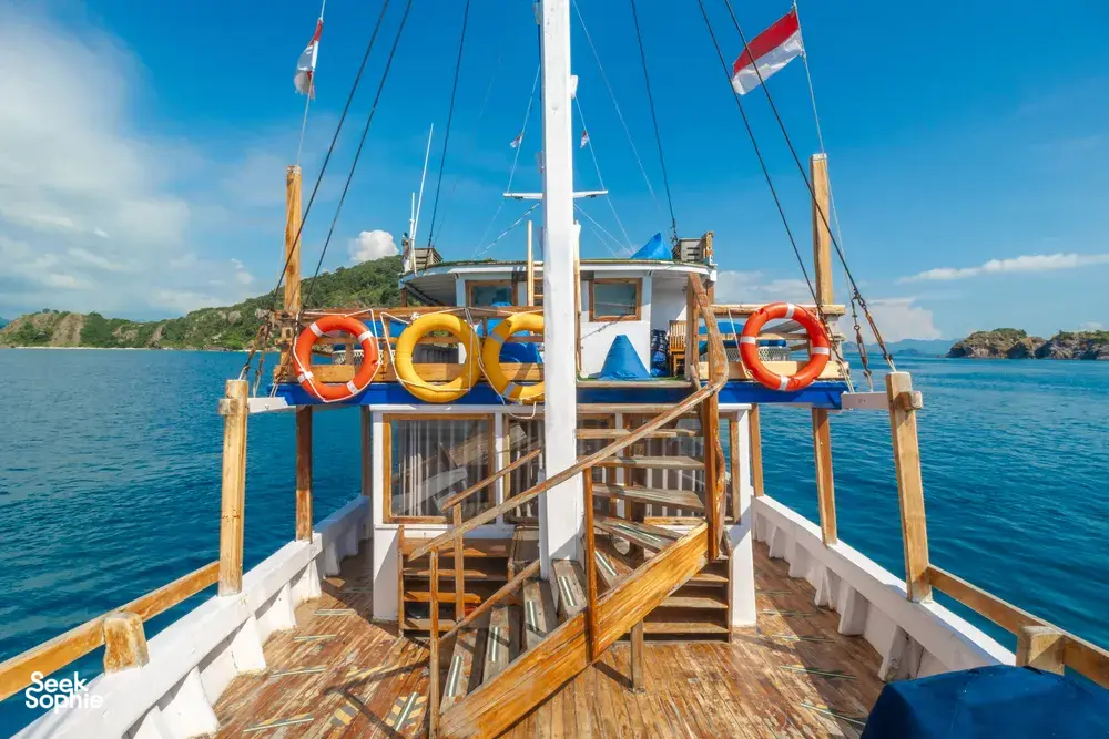 La Famille Komodo boat’s front deck with orange lifebuoy rings and sail flags, mast over open water on Labuan Bajo