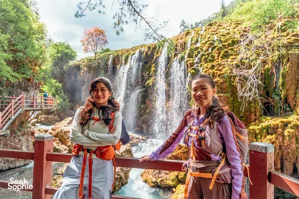 Two travellers pose at a wooden railing by a multi-tier waterfall near Shangri-La during a 6-day trek loop