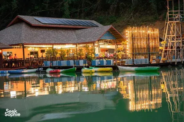 Lit pool and lodge buildings reflected in the night water, with colourful floats by the deck at Cheow Lan Lake