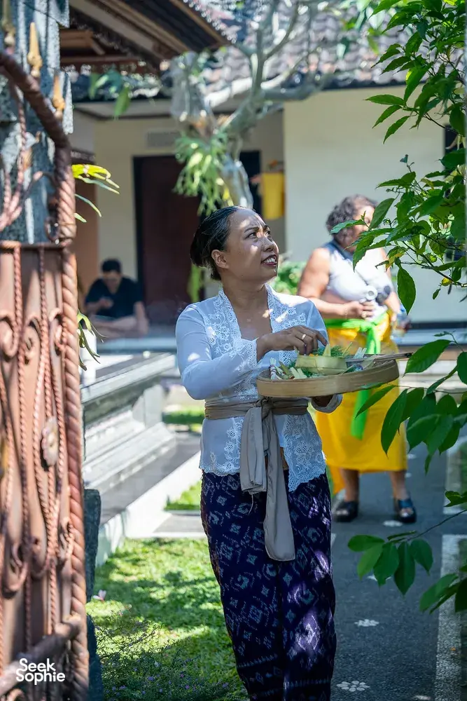 Balinese woman arranging canang sari offerings with flowers and incense in a garden courtyard, during a Ubud ceremony