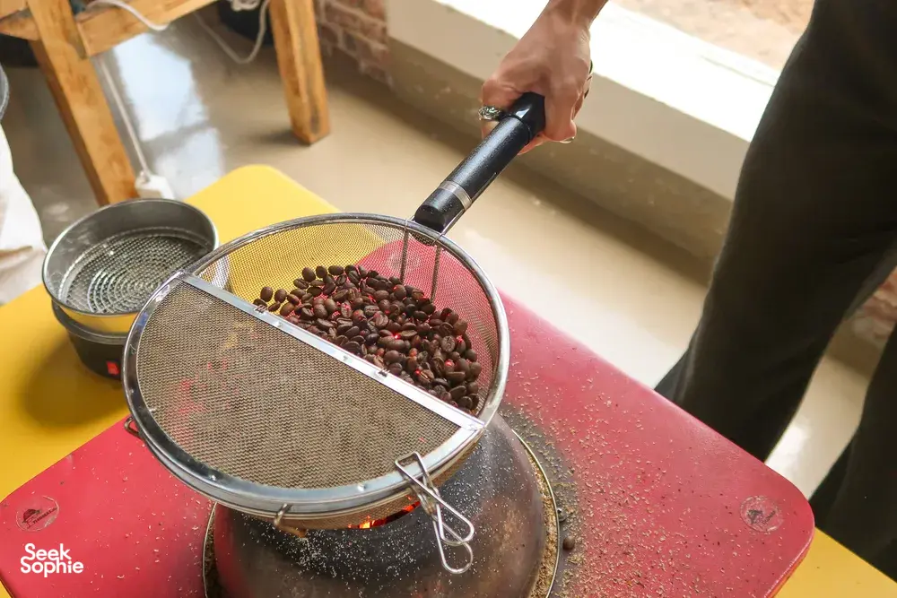 Seeker Yunnan Coffee Estate, Lijiang: close-up of hands sifting roasted coffee beans through mesh over a red roaster