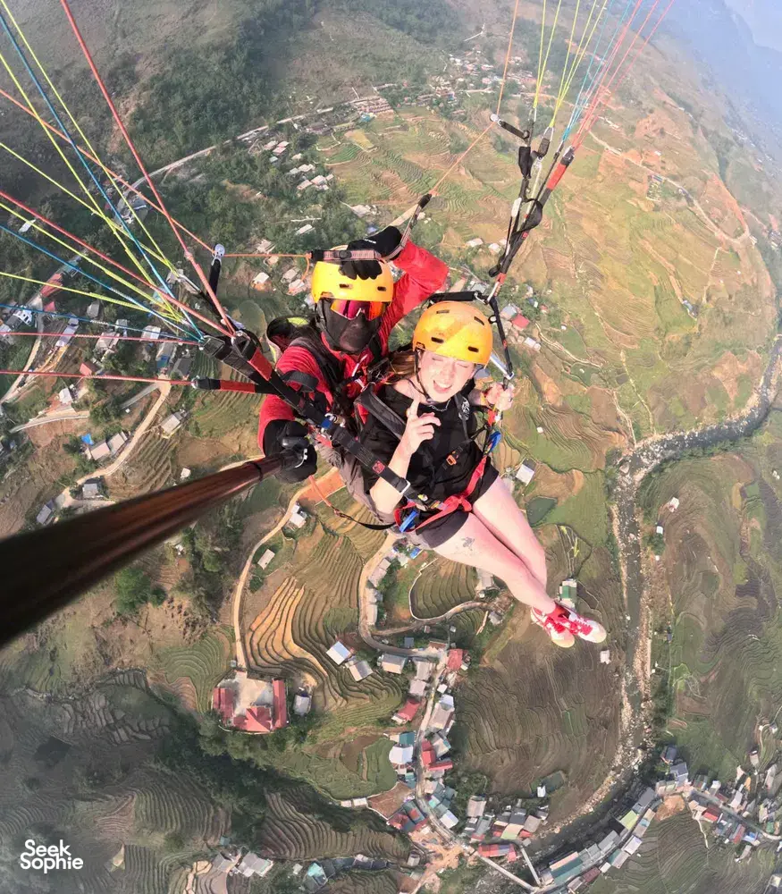 Tandem paragliding over stepped hillside rice terraces with two orange-helmeted riders holding the lines in mid-flight in Sapa