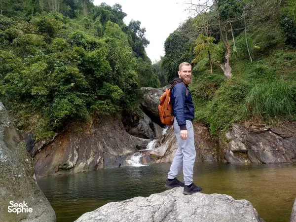 Solo trekker with backpack stands on a river rock beside a waterfall through lush scenery on the Ha Giang loop