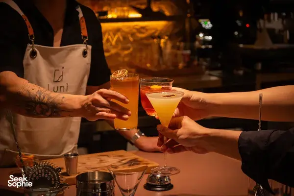 Hands raise two by-the-glass cocktails with citrus garnishes for a toast at an intimate Singapore cocktail bar.
