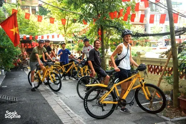 Group of riders on yellow bicycles on a quiet Hanoi lane under red-and-yellow flags, part of a Hidden Wartime cycling route