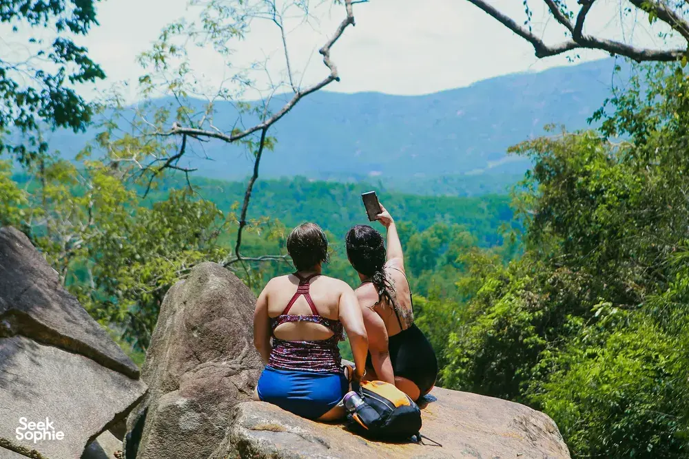 Backpackers seated on a boulder overlooking a lush valley in Hoi An, pausing for views during a waterfall motorbike adventure