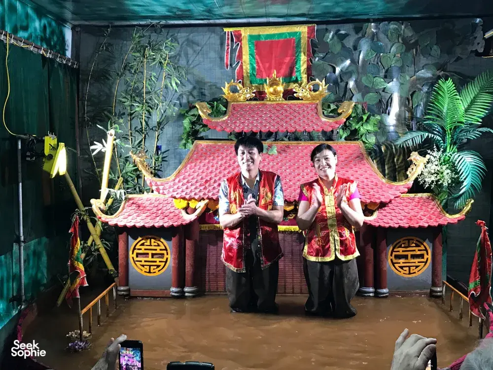 两位身穿红色戏服的第七代水上木偶艺人在河内传统戏台上表演,背景是装饰精美的寺庙风格布景