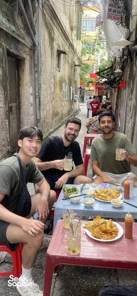 Travellers eating together at a roadside table in a narrow Hanoi lane, part of a beyond-Old-Quarter food adventure