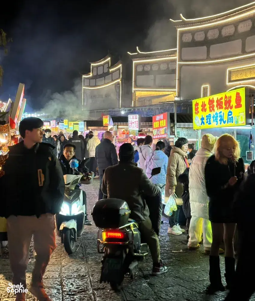 Lijiang night market street crowded with pedestrians and scooters under neon-lit traditional buildings, with misty smoke in the air