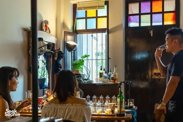 Guests seated at a wooden tasting table, swirling small-batch whisky in a hidden home bar in Katong, Singapore under candlelight