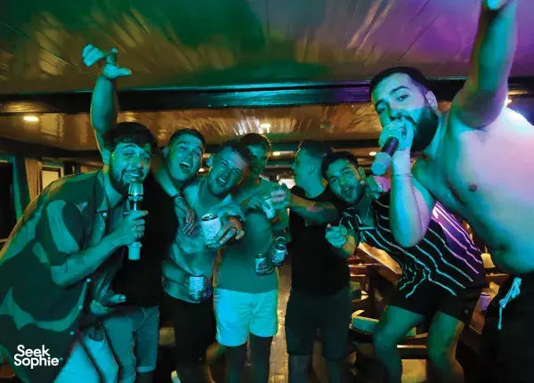 Group of friends cheering and drinking on board Oasis Bay Party Cruise at night under neon lighting, with drinks in hand in Halong Bay