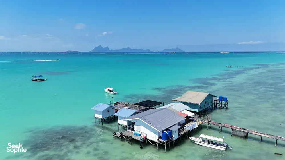 Pemandangan dari udara banglo atas air dan jeti selam di Semporna di atas laut biru firus yang jernih, dengan bot berlabuh dan bukit-bukau di kejauhan