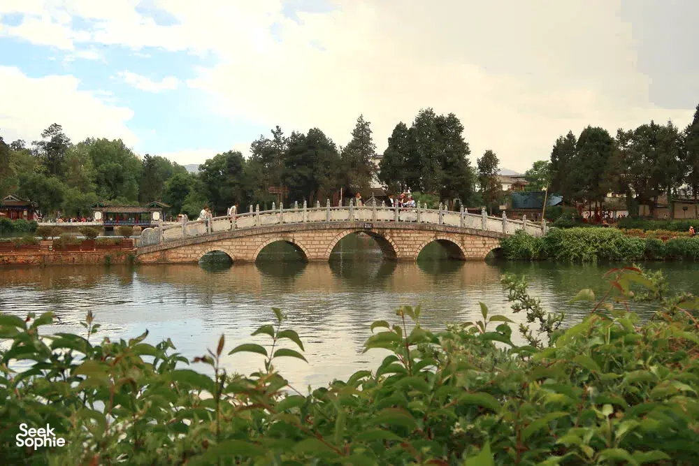 Stone arched bridge spanning a canal with lush trees in Lijiang Old Town during a local-guided Hidden Stories walk