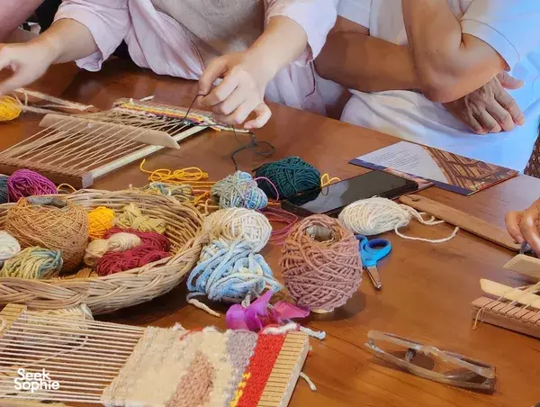 Hands working on miniature looms surrounded by neatly arranged yarn skeins during an art-and-crafts class in Thailand