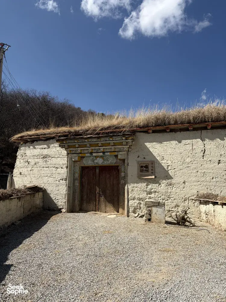 Whitewashed Tibetan outbuilding with a carved wooden door under blue sky, with tall reeds beside it in Shangri-La