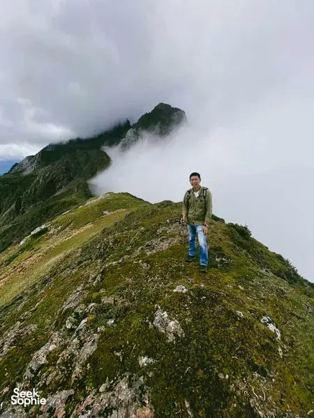 Hiker standing on a misty alpine ridge above the clouds, surrounded by green slopes, on a guided hike to Jade Dragon Snow Mountain in Lijiang