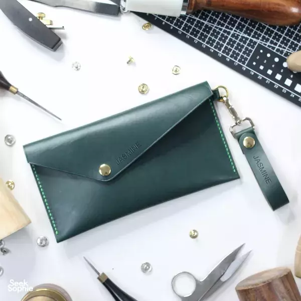 Dark green handmade clutch wallet marked “JASMINE” with brass snap on a workbench at Hides and Thread workshops in Singapore