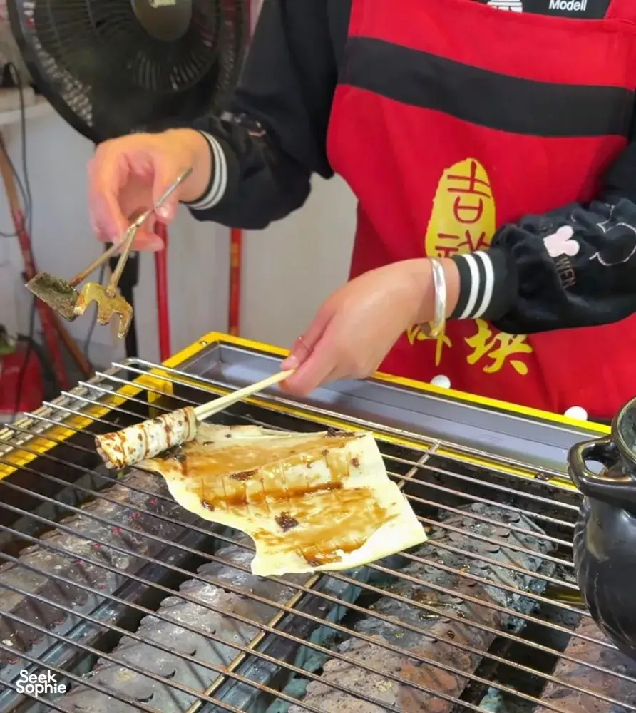Hands cook thin flatbread on a hot wire rack in Lijiang, with sauce and seasoning at a street food stall