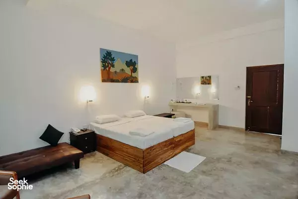 Sunlit guest bedroom with wooden bed, table seating, wall lamps and a door in the lodge in Anuradhapura