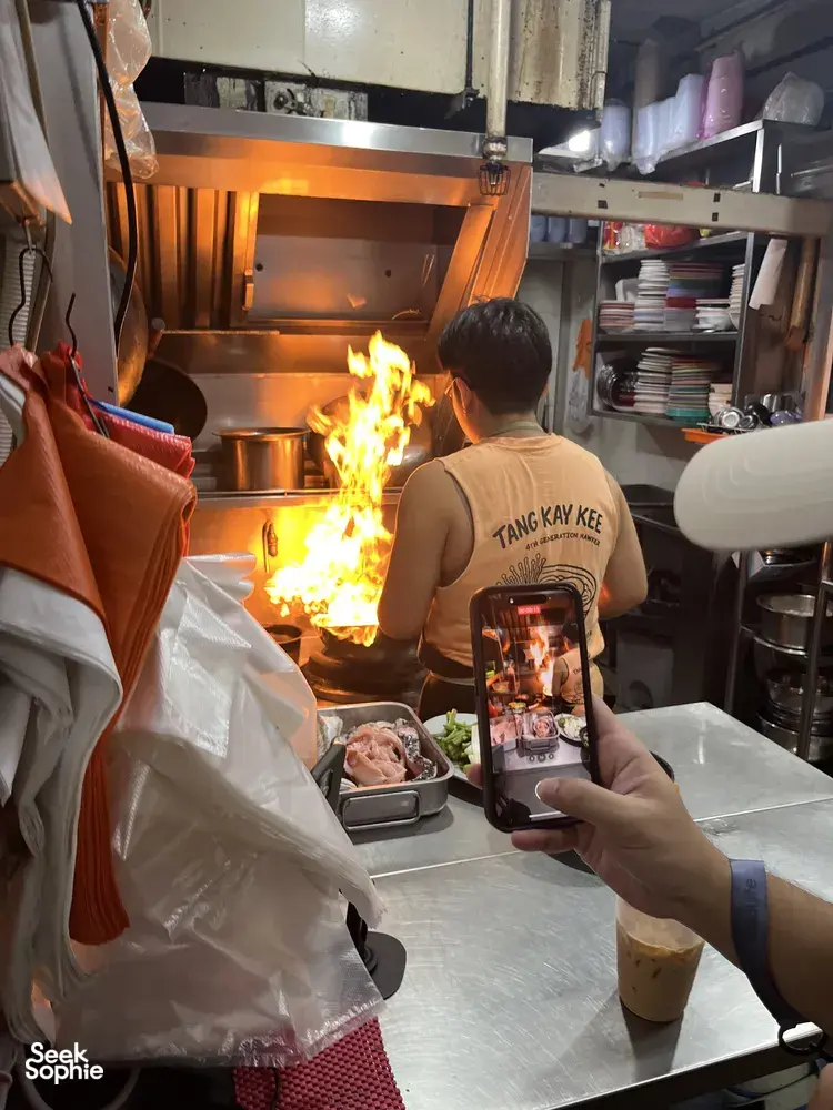 Seorang tukang masak mengendalikan wok yang sedang menyala di dapur gerai penjaja Singapura, sementara para pengunjung menonton dan mengambil gambar dengan telefon
