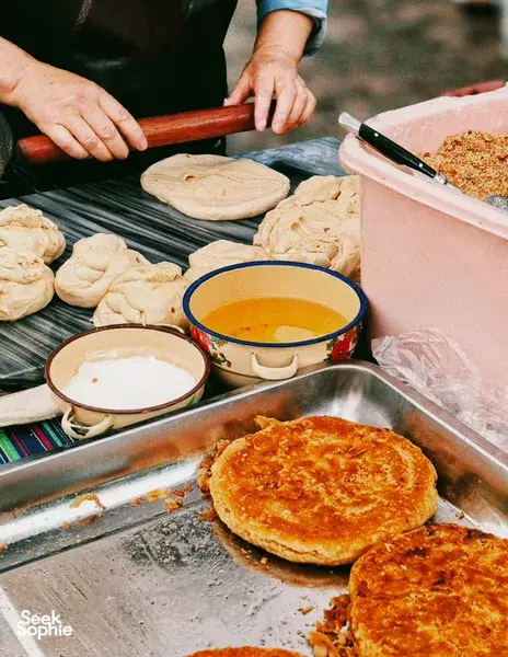 Hands roll and cook round flatbreads on a hot griddle with bowls of dipping sauces, part of a Lijiang food hunt