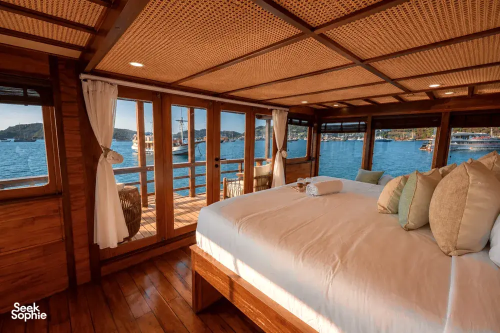 Phinisi cabin bedroom with a neatly made double bed, pillows and sheer curtains, looking out over the water in Labuan Bajo