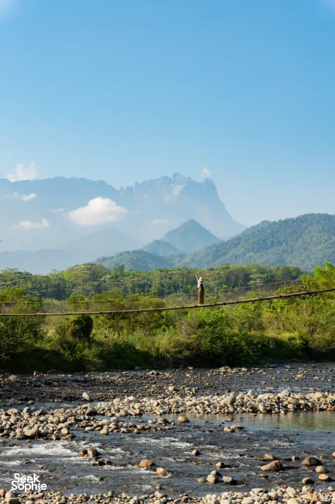 Hiker standing on a lookout above a river with a mountain horizon during a Borneo Overland Sabah road trip with Mt Kinabalu