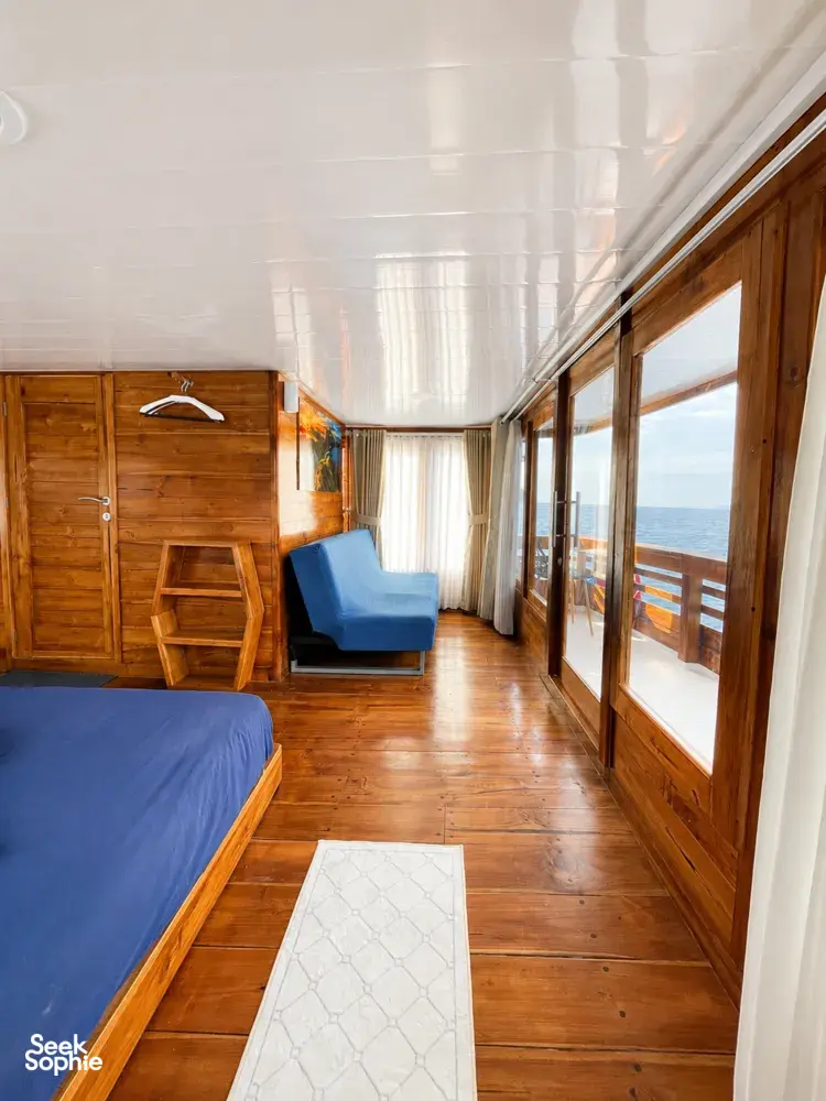 Wooden cabin interior with a blue sofa chair, bed and sea-facing windows, showing comfort for a 3D2N shared Komodo cruise, Komodo Island