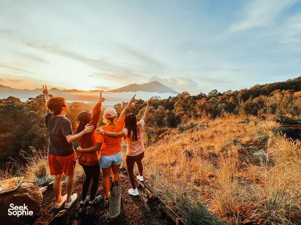 Group of travellers posing with arms raised during a sunset jeep stop on a grassy ridge above volcanic valleys in Bali, Mt Batur