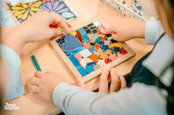 Hands arranging coloured ceramic mosaic tiles in a wooden frame at QMosaic Artspace & Workshop in Bat Trang, near Hanoi