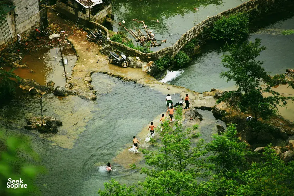 People wading in a shallow clear river in Cao Bang, with motorbikes parked along stone embankments and a relaxing crowd