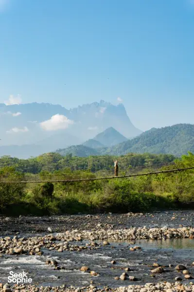 Hiker standing on a lookout above a river with a mountain horizon during a Borneo Overland Sabah road trip with Mt Kinabalu