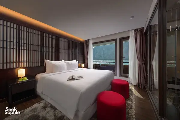 Oasis Bay Party Cruiser cabin with a made-up bed, red ottoman seating and sea-view windows overlooking karst scenery on the Halong Bay