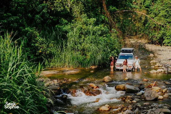 Borneo Overland group wading through a shallow rocky river beside off-road jeeps during wild Sabah camping, en route to Mt Kinabalu