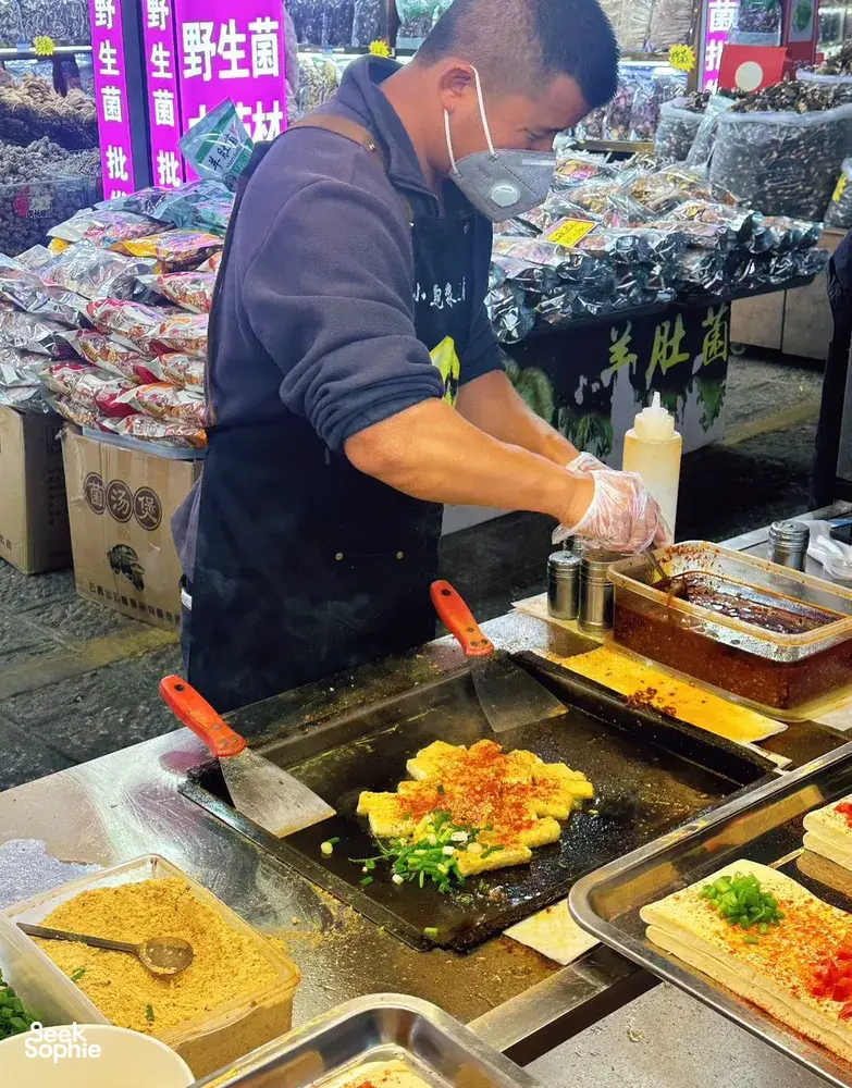 Vendor in a Lijiang night market grills savoury pancakes with chopped vegetables on a hot griddle, preparing servings for customers