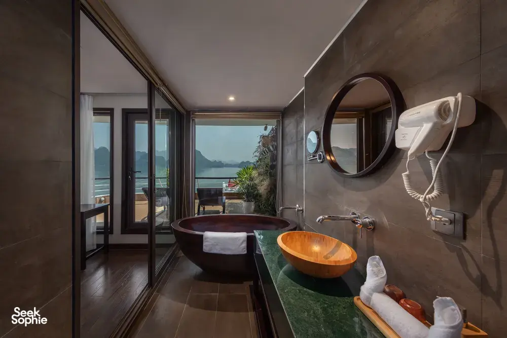 Bathroom on the Oasis Bay Party Cruiser with a stone bathtub and round mirror, looking out over the water and hills of Halong Bay