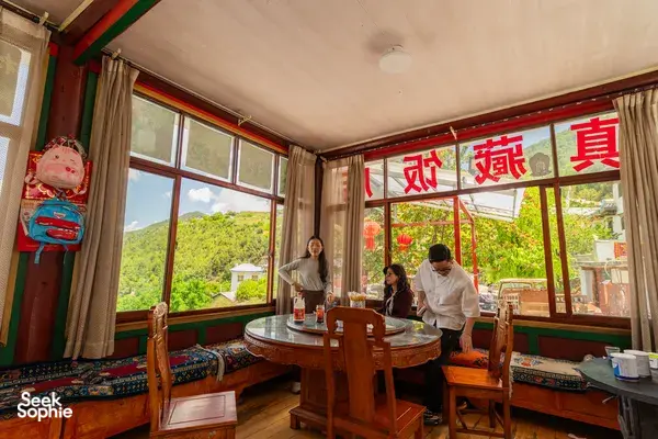 Lodge dining room in Shangri-La with large windows to green hills, travellers at a round table preparing snacks