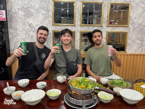 Para pengembara duduk di meja restoran dengan hot pot untuk dikongsi bersama dan mangkuk-mangkuk kecil, menikmati pengembaraan makanan Hanoi dalam kumpulan kecil