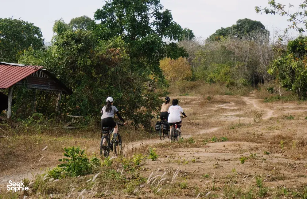 Cyclists ride on a sandy track through Langkawi countryside, passing a small roadside house and dense greenery