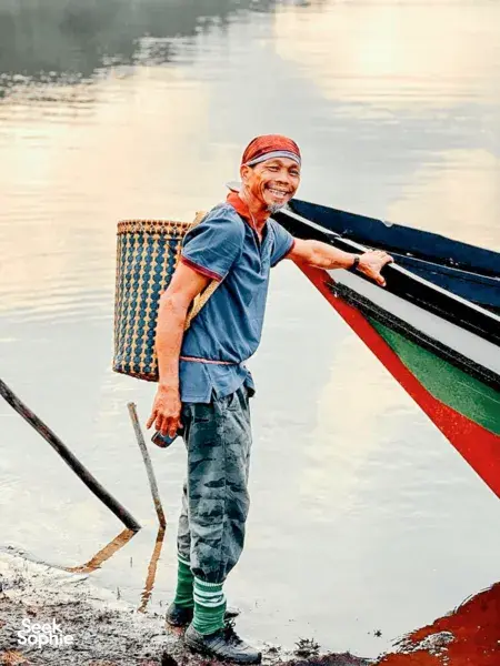 Smiling man standing at the edge of a longboat on a calm river near Kuching, wearing a headscarf as dawn light reflects