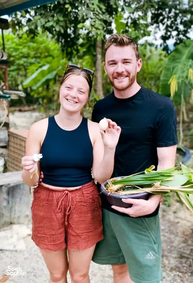 Two Kebun Lidstari hosts smiling in a lush Langkawi permaculture garden, holding fresh greens and a serving bowl for cooking.