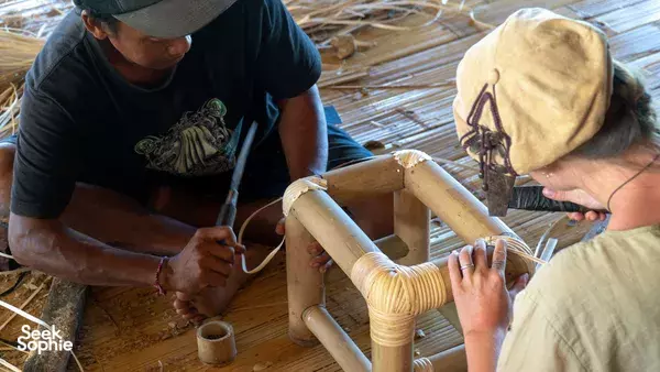 Indonesia woodworking class: two people cut and smooth bamboo pieces as the tutor guides them at a woven-mat workspace