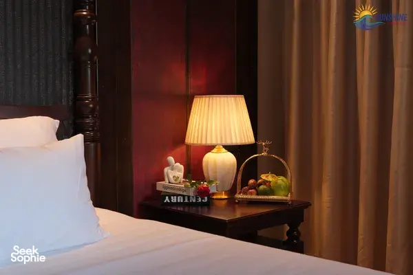 Sunshine Golden Cruises cabin bedside table with an illuminated lamp, fruit tray and bundled bedside linens against dark wood walls