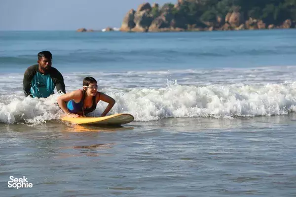 Beginner surfer paddling on a board with an instructor in the shallows at Weligama beach as waves roll in