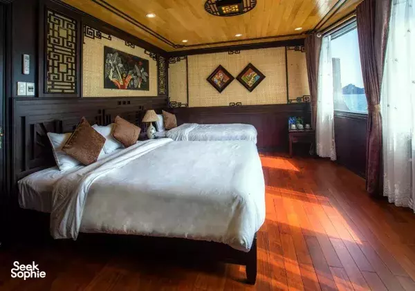 Wooden cruise cabin bed with pillows and floor-length curtains, decorative wall art, and a window with sea light in Cat Ba