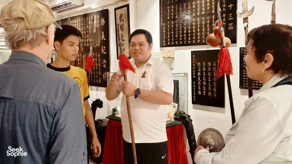 A guide in a white polo explains a lion dance instrument while guests stand around red drapes and calligraphy in Ho Chi Minh City
