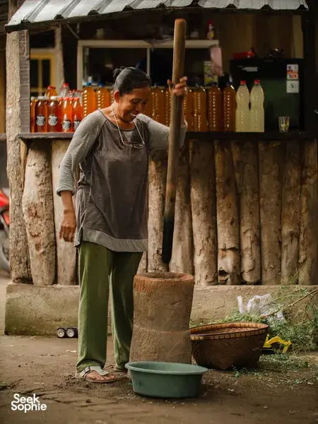A man prepares drinks at an outdoor village counter in Lombok, with rows of amber bottles and a serving cart behind him