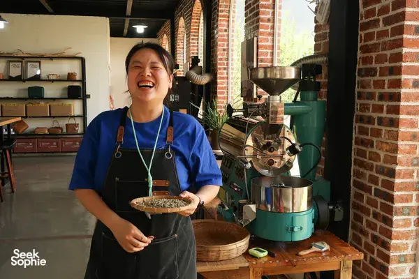 Bengkel kopi istimewa dipandu hos di Seeker Yunnan Coffee Estate, Lijiang: seorang wanita memegang dulang berisi biji kopi panggang di sebelah mesin pemanggang